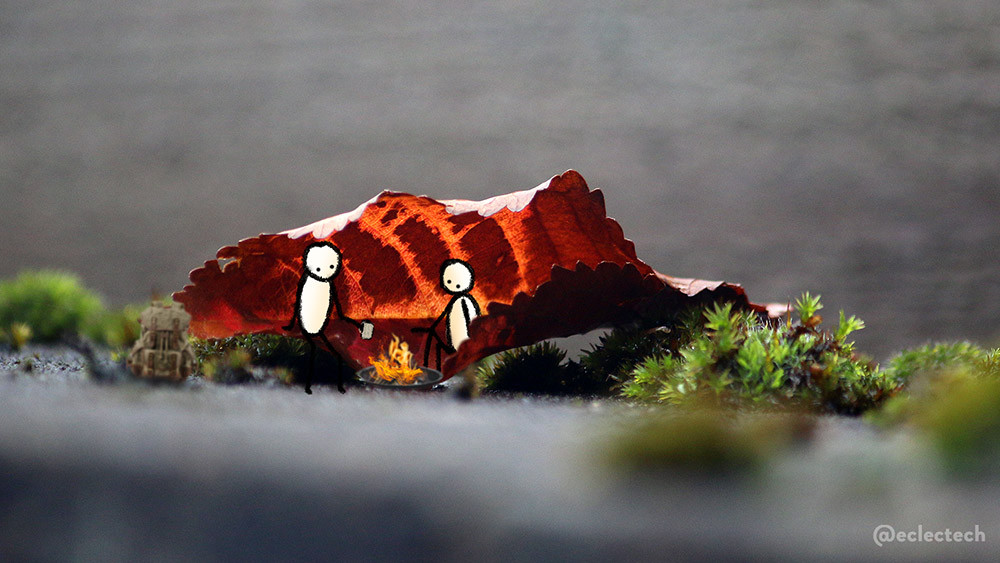 A photo of a curled up autumn leaf resting on a wall with mossy clumps either side. The leaf has dark and bright orange stripes, and is the focus of the photo, with the surround of dark green of moss and grey wall in the fore and background slightly blurred. I have drawn two simple figures sitting in the shelter of the leaf, either side of a small camp fire. One holds a cup of something hot. A dark green rucksack rests on the ground to one side of them.