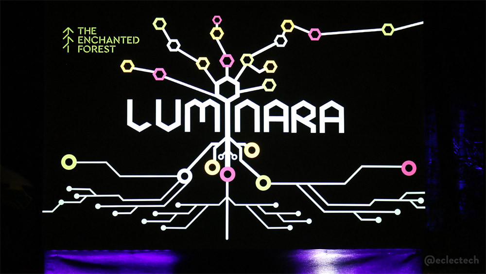 A photo of the illuminated show name sig. In the top left is their tree-like logo with THE ENCHANTED FOREST alongside. In bright white letters in the centre is the show name LUMINARA, and a design where the top reflecting circuits and connections; organic ones at the top, and circuit board tech ones at the bottom.The background is black, with some purple light along the bottom.