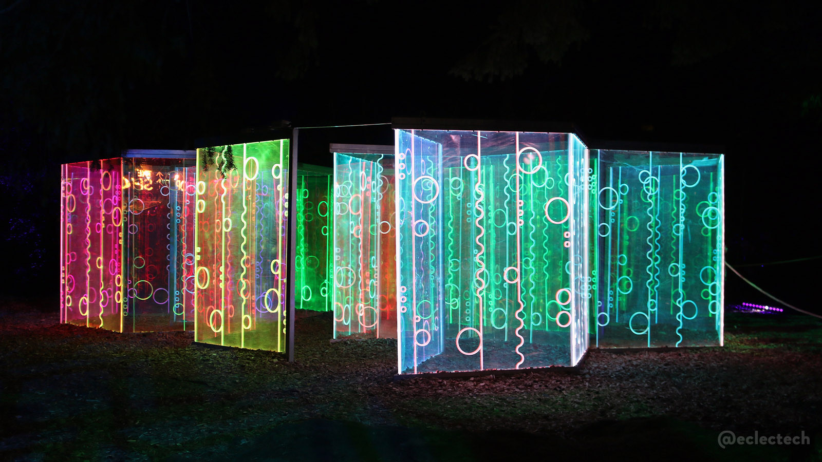 Photo of the Living Networks maze form the 2025 Enchanted Forest, taken from outside the maze. Against a black backdrop are a series of tall panels decorated with circles, and vertical squiggly lines, lit up in a variety of colours, from bright pinks, to yellow, blues and greens. The panels are maybe a couple of metres tall, and there are openings where people can walk through.