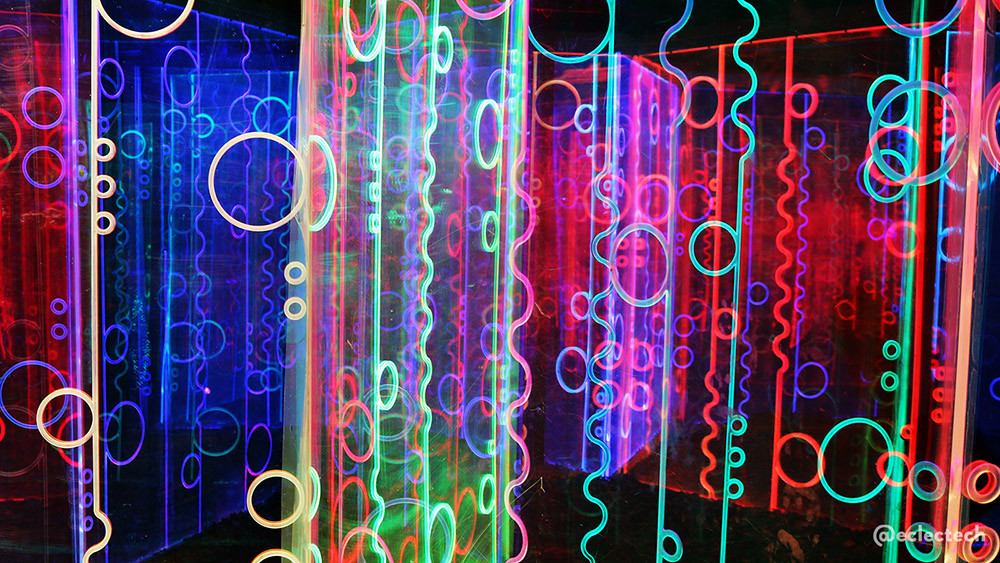 Photo of the Living Networks maze from the 2025 Enchanted Forest, taken from outside the maze, but close up. The frame is filled with a series of tall panels decorated with circles, and vertical squiggly lines, lit up in a variety of colours, from bright pinks, to dark blue and greens, all appearing to overlap and merge.