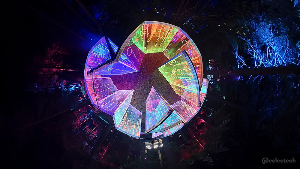 Photo of the Living Networks maze from the 2025 Enchanted Forest, taken from the centre of the maze with a 360 degree camera, held high above the panels. The surround is black, with some illuminated trees, and the maze forms an almost circle of light, with a zigzag shape in the middle - the path running through it.