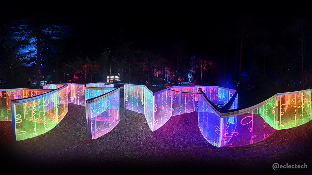 Photo of the Living Networks maze from the 2025 Enchanted Forest, taken from the centre with a 360 camera held high above the panels. The photo is shown in a slightly edited equirectangular projection, so the panels of coloured squiggles, circles and lights are spread our horizontally across the photo, with some illuminated trees behind.