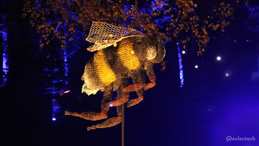 A photo of a large model of a bee, made of wire netting, and brightly illuminated against a dark blue backdrop with a hint of trees.