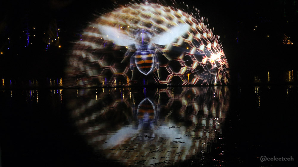 A huge light projection of a striped bee on a honeycomb semi circle, also reflected in the water of the loch.