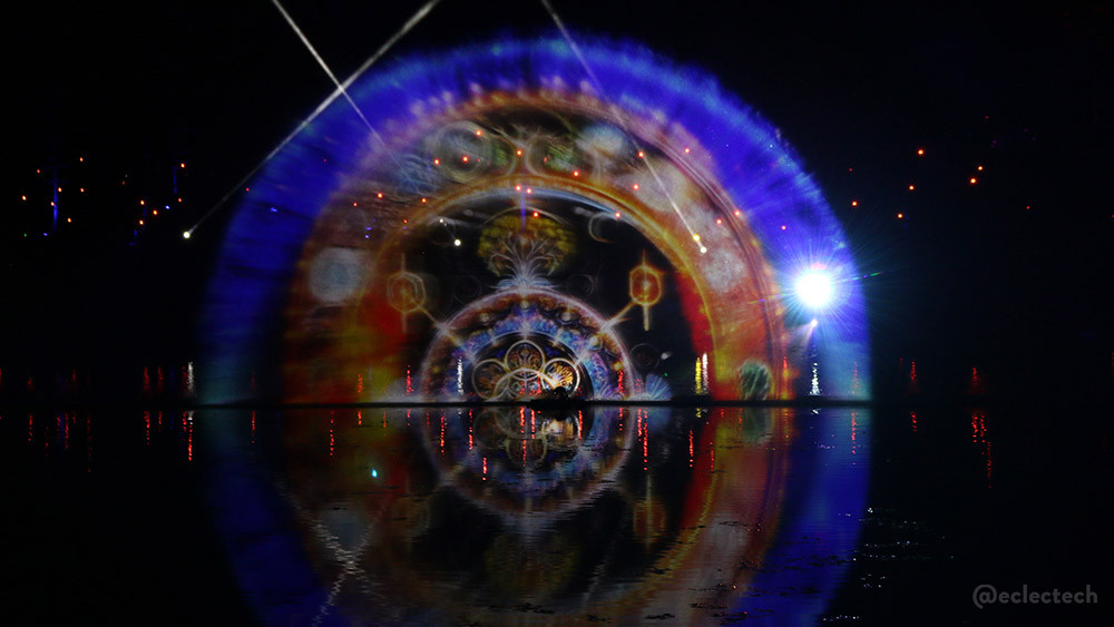A semi circle of projected pattern over a loch with a faint reflection in the water. The outer ring is deep blue with predominantly red and yellow detailed patterns in the smaller rings.