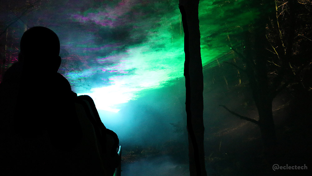 A shimmering green and pink aurora type light spread across the pathway, with a silhouette of some people on the left, and a tree trunk near the centre.