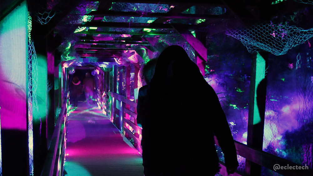 A view down a wooden bridge. There is the silhouette of a person in the foreground, and the bridge is a wooden slatted tunnel, lit up in pinks and pale green.