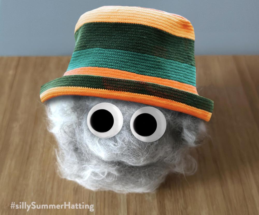 Original alt txt: a rather large, roughly ball-shaped pile of pale grey and white floof. it's on a wooden table mat and in front of a blue background.
My addition: The floof now has large googly eyes, above a smiling mouth naturally formed in the ball, that I have only very slightly emphasised. It wears a knitted hat in orange and green stripes. It looks super cheerful.